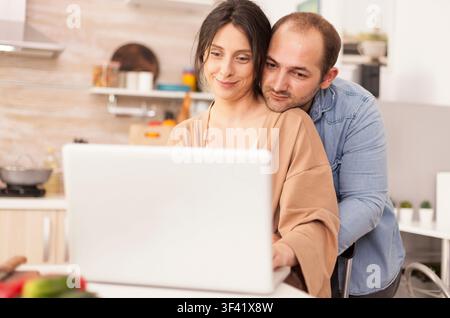 Lächelndes Paar verwendet Laptop in der Küche, Mann umarmt Frau, verbindet Laptop-Arbeit, Computerarbeit und Fernarbeit, während des Frühstücks in einer Küchenroutine zu Hause. Stockfoto