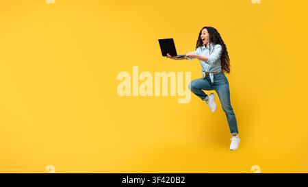 Aufgeregte junge Frau springt mit Laptop vor hellgelbem Hintergrund, Geschäftskonzept Stockfoto