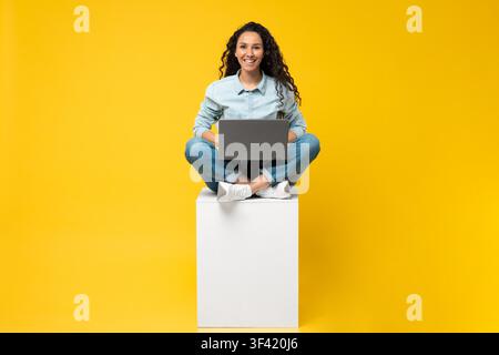 Lächelnde junge Frau, die einen Laptop benutzt, sitzt mit gekreuzten Beinen auf einem weißen Würfel vor gelbem Hintergrund Stockfoto