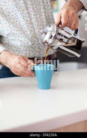 Älterer Mann macht Kaffee mit French Press, betont den älteren Mann, der während des Frühstücks in der Küche Kaffee mit French Press zubereitet und ihn in eine blaue Tasse gießt, alter Mensch. Stockfoto