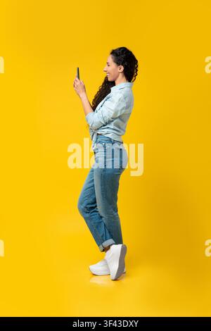 Junge Frau verwendet Smartphone im Profilblick, stehend auf gelbem Hintergrund Stockfoto