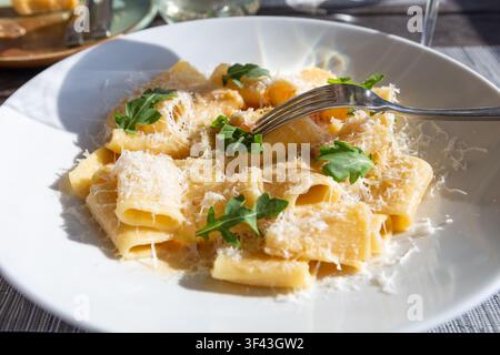 Kremiges Rigatoni-Pasta-Gericht serviert mit geriebenem Parmesan und grünen Rucolablättern auf einem weißen Teller mit Gabel. Stockfoto