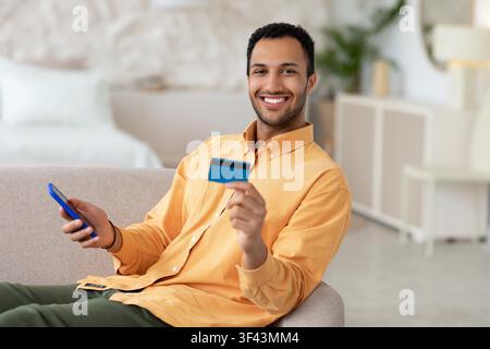 Lächelnder junger schwarzer Mann, der zu Hause eine Kreditkarte und ein Smartphone hält, Online-Shopping-Konzept Stockfoto