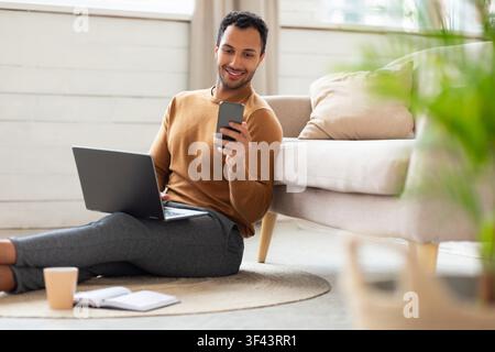 Lächelnder junger Mann, der einen Laptop und ein Smartphone benutzt, während er zu Hause auf dem Boden sitzt, Geschäftskonzept Stockfoto