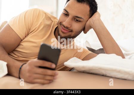 Lächelnder junger schwarzer Mann liegt auf dem Sofa und verwendet ein Smartphone zu Hause, entspannte Stimmung Stockfoto
