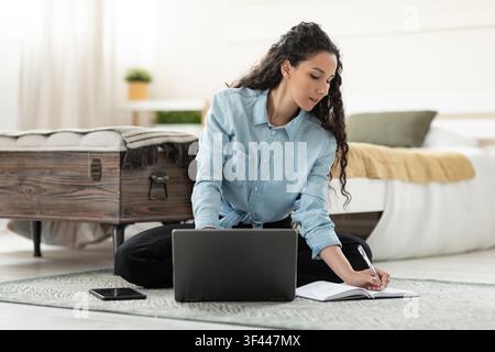 Konzentrierte junge Frau gemischter Herkunft arbeitet an einem Laptop und macht Notizen im Homeoffice Stockfoto