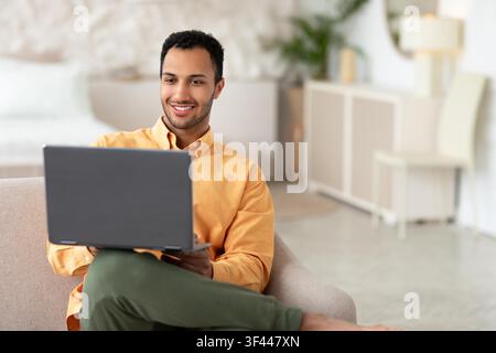 Lächelnder junger Mann gemischter Herkunft, der zu Hause einen Laptop nutzt, Home-Office-Konzept Stockfoto