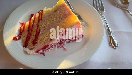 Ein angerichtetes Stück Himbeer-Kuchen mit Sahneglasur und roter Beerentropfengelee, serviert mit einer Gabel auf einer weißen Tischdecke. Nahaufnahme des Essens Stockfoto
