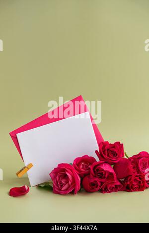 Leere Grußkarte mit passendem Umschlag, der mit einer Holzklammer befestigt ist, neben lebendigen rosa Rosen auf einem weichen Pastellhintergrund angezeigt. Blumenarrangement mit großzügigem Kopfraum schafft eine elegante Komposition für Einladungen, Glückwünsche, Feiern, Hochzeiten, Geburtstage, Jubiläen, Valentinstag, Muttertag oder romantische Nachrichten. Stockfoto