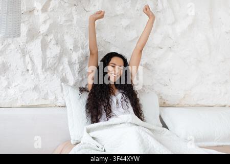 Glückliche junge lateinamerikanische Frau, die sich im Bett streckt, Morgenroutine, Entspannungskonzept Stockfoto