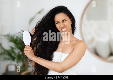 Frustrierte Frau mit langen dunklen lockigen Haaren, die zu Hause ihre Haare bürstet, Selbstpflege-Konzept Stockfoto