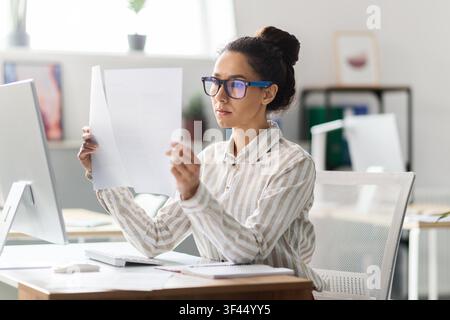 Konzentrierte junge asiatische Frau, die Dokumente an einem modernen Bürotisch durchsieht, Geschäftskonzept Stockfoto