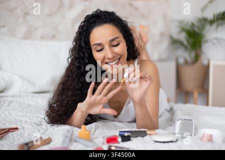 Zufriedene junge Frau, die Make-up mit Beauty-Produkten auf dem Bett auftragt, häusliche Umgebung Stockfoto