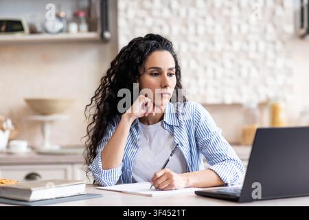 Konzentrierte hispanische Frau, die Notizen macht, während sie zu Hause am Laptop arbeitet, Geschäftskonzept Stockfoto