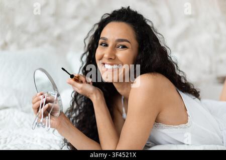 Lächelnde junge Frau mit lockigen Haaren, die Make-up mit einem Spiegel im Schlafzimmer aufträgt, Hautpflegekonzept Stockfoto