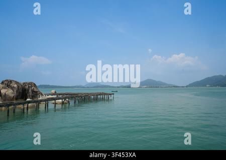 Eine ruhige tropische Küstenlandschaft der Insel Pangkor mit einem verwitterten Pier, der sich in ruhige türkisfarbene Gewässer erstreckt, flankiert von großen Granitfelsen. Stockfoto