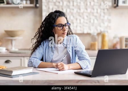 Konzentrierte hispanische Frau, die Notizen macht, während sie im Home Office an einem Laptop arbeitet Stockfoto