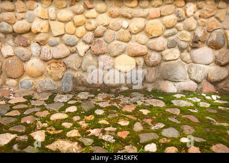 Foto mit Kopfsteinpflaster-Wand, grüner Moos-Bodenhintergrund. Alte rustikale Steinmauerstruktur mit texturierten runden Felsbrocken und altem Kopfsteinpflaster-Boden Stockfoto