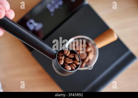 Geröstete Kaffeebohnen, die mit einem Messlöffel und einem Glasbehälter auf einer digitalen Waage gemessen werden, bevor sie gemahlen und der Kaffee aufgebrüht wird. Stockfoto