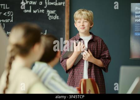 Kaukasischer Schuljunge präsentiert ein Codierungsprojekt und hält ein Tablet im Klassenzimmer, Klassenkameraden beobachten ihn in der Nähe der Tafel. Nützlich für MINT-Bildung, Schult Technologie, Schülerlernkampagnen Stockfoto