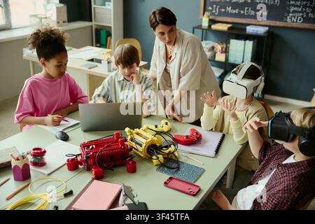 Breite Aufnahme von vielfältigen Schulkindern, die Laptop, Robotik-Kit und Virtual-Reality-Headsets nutzen, mit einer Lehrerin, die den Unterricht in STEM-Fächern leitet. Ideal für die Förderung von Bildungstechnologie Stockfoto