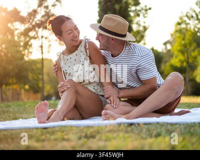 Ein Paar sitzt eng aneinander auf einer Picknickdecke und teilt einen liebevollen Moment im warmen Schein der untergehenden Sonne. Stockfoto