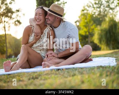 In goldenem Abendlicht gebadet, genießt ein glückliches Paar ein romantisches Treffen im Freien auf einer Picknickdecke. Stockfoto