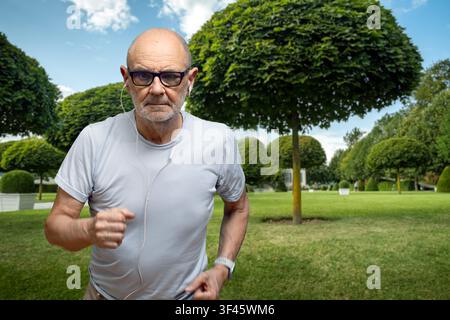 Senior joggt durch einen gepflegten Park, trägt Ohrstöpsel und eine Brille, konzentriert auf Outdoor-Fitness, gesunden Lebensstil und aktives Altern und genießt dabei Stockfoto