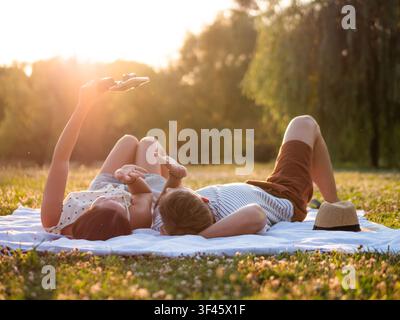 Ein junges Paar entspannt sich auf einer Picknickdecke in einem friedlichen Park, während die Sonne untergeht. Seite an Seite liegend genießen sie das warme Licht der Goldenen Stunde, wäh Stockfoto