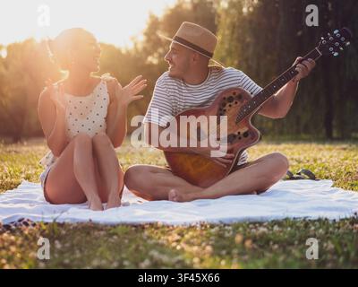 Ein junges Paar umarmt den Zauber der goldenen Stunde mit einer Outdoor-Jam-Session in einem malerischen Park. Auf einer Picknickdecke bequem sitzend, genießen sie liv Stockfoto