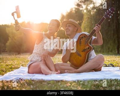 Ein fröhliches Paar genießt ein entspanntes Picknick im Park während der goldenen Stunde und macht ein Selfie mit einem Smartphone, das auf einem Hand-Held-Gimbal montiert ist. Der Mann hält Stockfoto