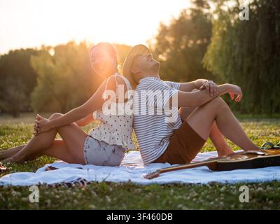 In warmem Abendlicht gebadet, genießt ein lächelndes Paar ein entspanntes Picknick im Freien, Rücken an Rücken sitzend. Stockfoto