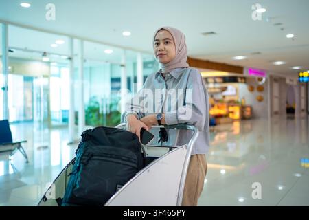 Nahaufnahme-Porträt einer jungen muslimischen Frau mit Gepäckwagen im Flughafenterminal, Reiselifestyle. Stockfoto
