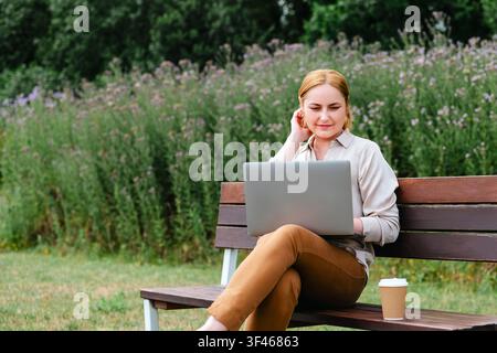 Frau arbeitet an einem Laptop, sitzt auf einer Parkbank mit Kaffee Stockfoto
