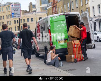 Ein DPD-Lieferfahrer lädt Pakete von einem Kurierwagen auf einer Straße in der City of London, UK, ab, während zwei Personen im Vordergrund auf der Bordsteinkante sitzen und eine Zigarettenpause machen Stockfoto