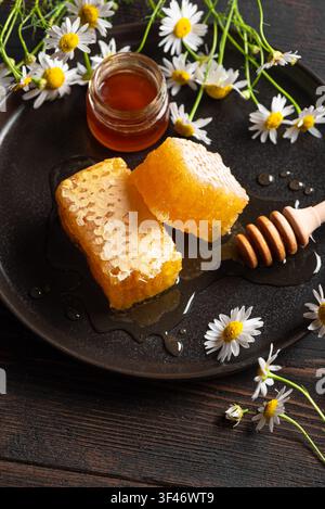 Goldener Honigwaben auf schwarzem Teller mit Kamillenblüten auf Holzunterlage, Kopierraum. Frischer Honig mit Wildblumen-Design, Honigernte-Konzept Stockfoto