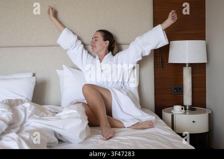 Junge Frau streckt ihre Arme, während sie in einem Bademantel auf einem Hotelbett sitzt Stockfoto