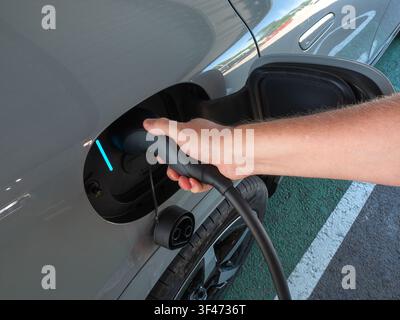 Nahaufnahme einer Person, die ein EV-Ladekabel in die Steckdose eines modernen grauen Elektroautos steckt. Eine beleuchtete zyanfarbene Statusanzeige lig Stockfoto