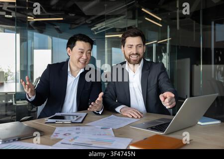 Verschiedene Geschäftspartner lächeln, diskutieren Daten und zeigen auf einen Laptop-Bildschirm während eines erfolgreichen Meetings in einem modernen Büro, was Teamwork und positive Zusammenarbeit symbolisiert Stockfoto