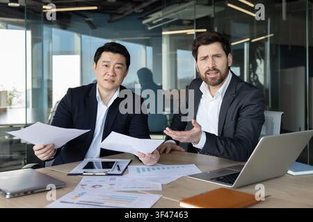 Zwei vielfältige Geschäftskollegen sitzen an einem Bürotisch, halten Papiere und drücken Verwirrung und Stress aus, während sie Finanzdokumente analysieren und eine schwierige Situation besprechen Stockfoto