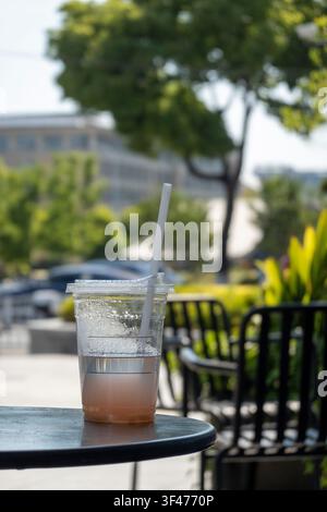 Ästhetische Außenszene mit einem klaren Plastikbecher mit einem Papierstrohhalm und Kondenswasser auf einem schwarzen Tisch. Ein üppiger grüner Bokeh-Hintergrund fängt eine Summe ein Stockfoto