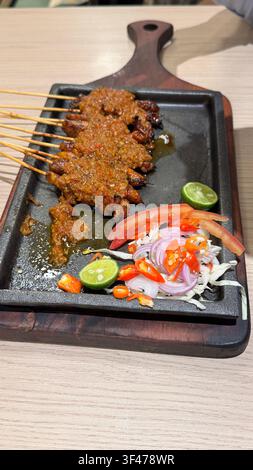 Indonesische Hähnchen-Satay-Spieße serviert mit Erdnusssoße, Chili, Limette, Tomate und geschnittener Zwiebel auf einem brutzelnden Teller. Stockfoto