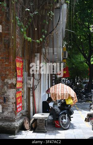 Vietnamesischer Verkäufer sitzt auf der Straße unter einem farbenfrohen Sonnenschirm im alten Viertel von Hanoi, Vietnam Stockfoto