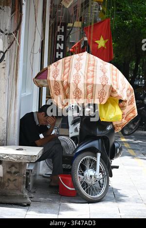 Vietnamesischer Verkäufer sitzt auf der Straße unter einem farbenfrohen Sonnenschirm im alten Viertel von Hanoi, Vietnam Stockfoto