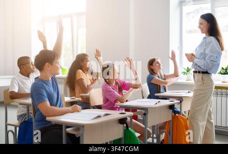 Verschiedene Schulkinder heben die Hände, um einem lächelnden Lehrer während einer interaktiven Unterrichtsstunde zu antworten Stockfoto