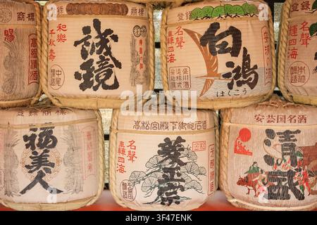 Gestapelte große traditionelle mit Stroh umwickelte Sakefässer mit handbemalten Kanji-Markennamen vor einem Schrein, Miyajima-Insel, Hiroshima, Japan Stockfoto
