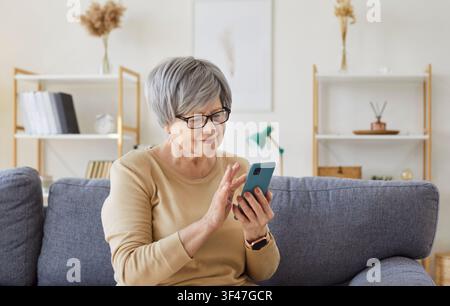 Seniorin sitzt auf der Couch im Wohnzimmer zu Hause und hält ein Mobiltelefon in den Händen. Stockfoto