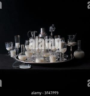 Antikes Kristall- und Pyrex-Laborglas mit Milch gefüllt, auf einem Silbertablett, um den Kosten von Glasgeräten gleichzusetzen, um die Kosten für die Ausstattung eines Labors zu veranschaulichen. Stockfoto