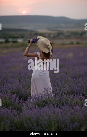 Frau in Sommerkleid mit Strohhut steht in einem blühenden Lavendelfeld während eines malerischen Sonnenuntergangs Stockfoto