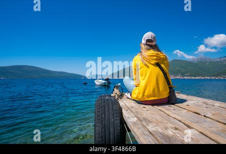 Junge kaukasische Frau mit langen blonden Haaren, die eine gelbe Jacke und eine Kappe trägt, sitzt auf einem hölzernen Steg am Wasser und schaut auf Boote in der Ferne. Stockfoto